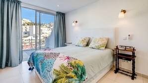 A bedroom with a large bed, a nightstand, a lamp, and a view of the city through a window. - Jacks Beach House Jack s Beach House Steps From (Lagoa)