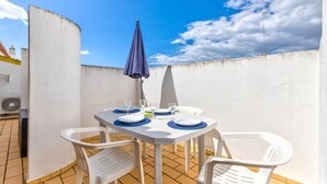 A terrace with a table set for two, white plastic chairs, a blue umbrella, and a white wall in the background. - 135 Monte Dourado Two Bed Apartment With Communa (Lagoa)