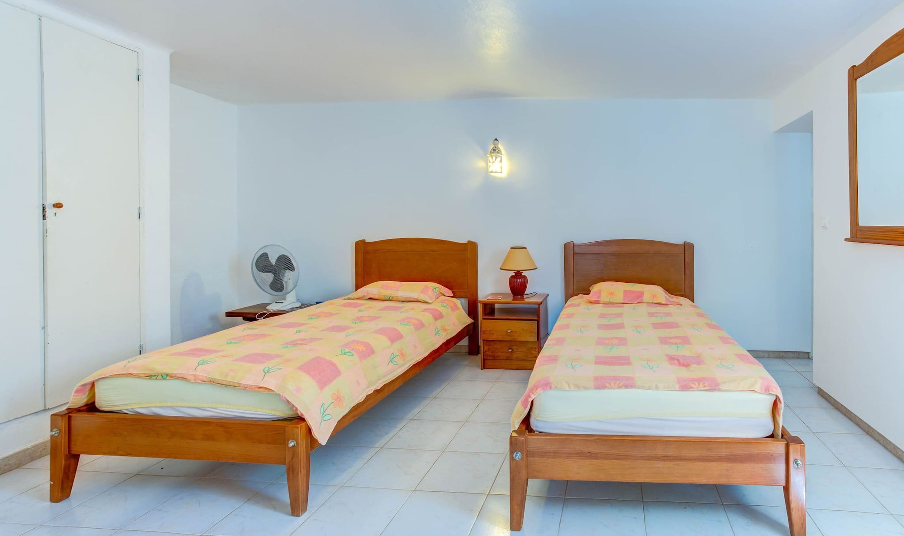 Una habitación con dos camas de madera, una mesita de noche con lámpara, un ventilador y una lámpara de pared.