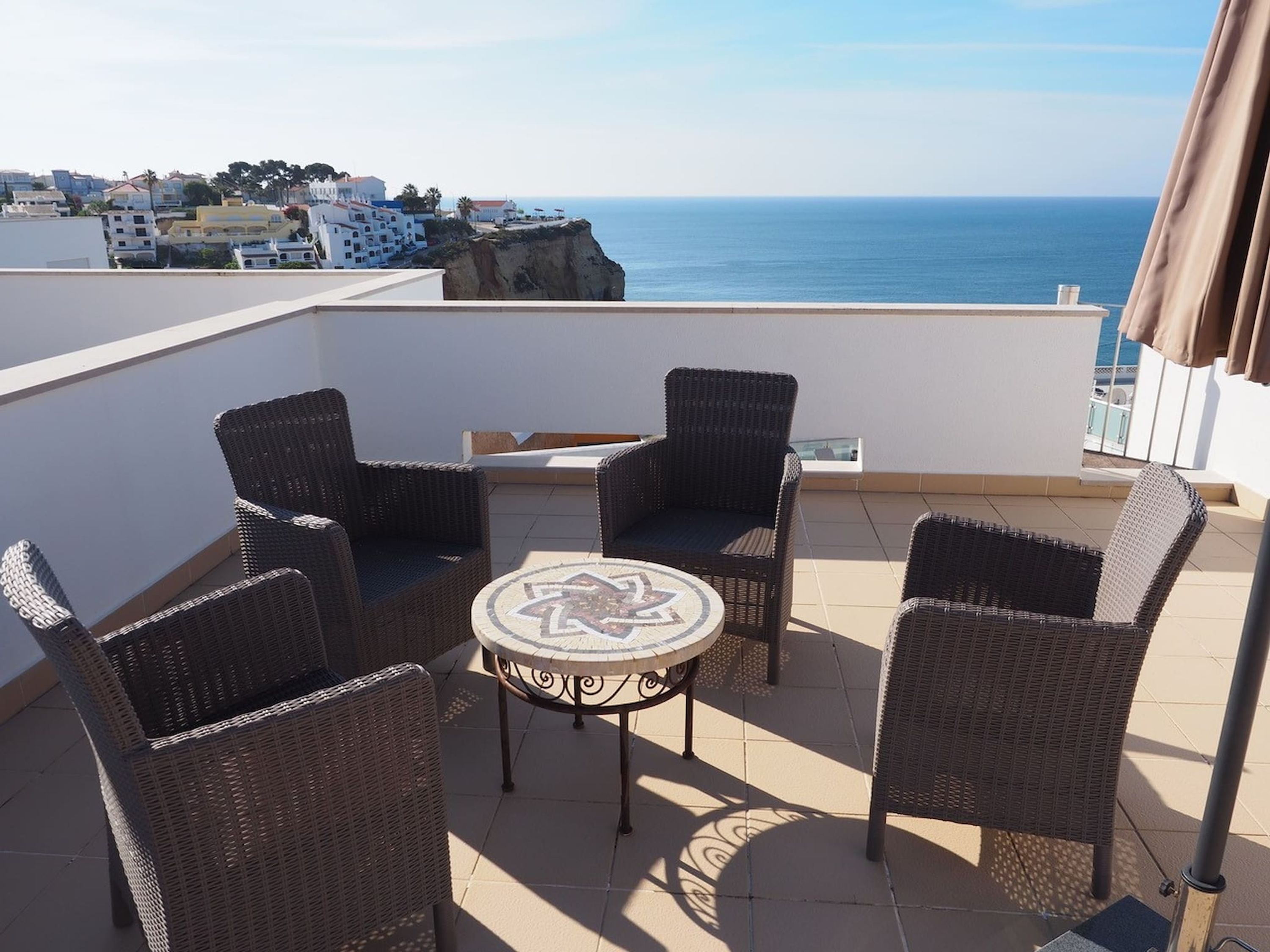 Una terraza con mobiliario de mimbre y una mesa, con vistas a un pueblo costero y al mar.