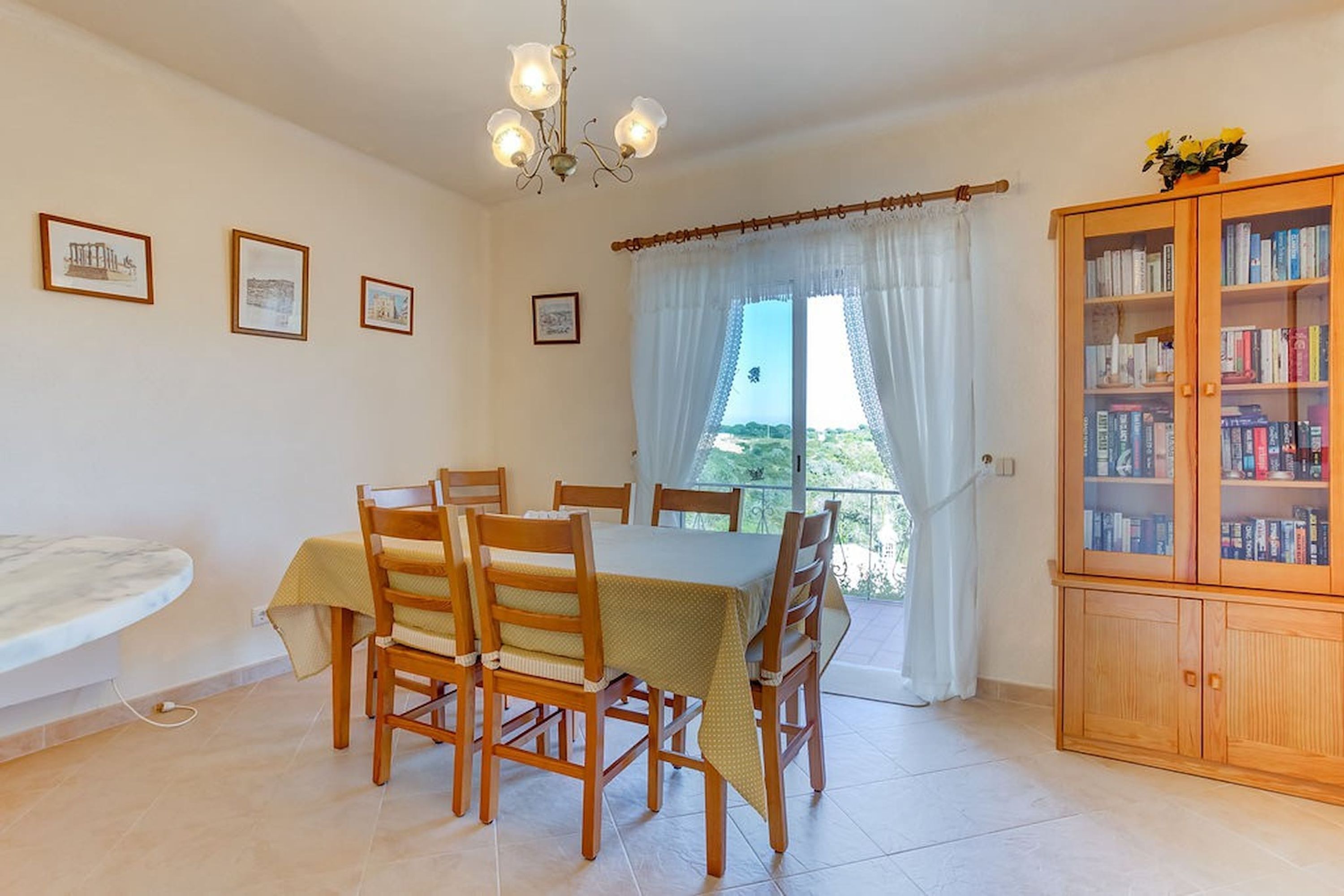 A dining area with a table, chairs, a chandelier, a bookshelf, and a window with curtains.