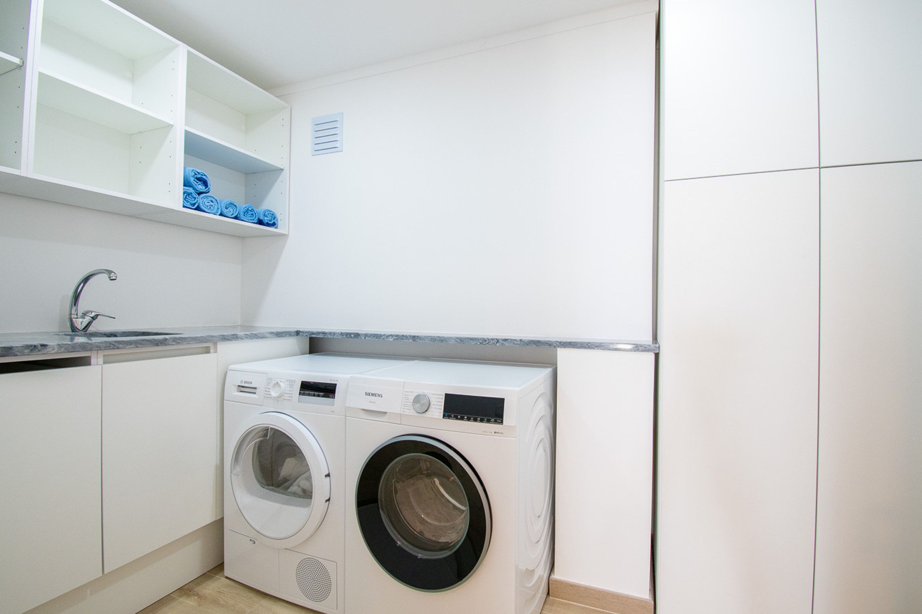 A compact laundry area with a front-loading washing machine, a sink, and a shelf with folded towels.