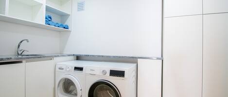 A compact laundry area with a front-loading washing machine, a sink, and a shelf with folded towels.
