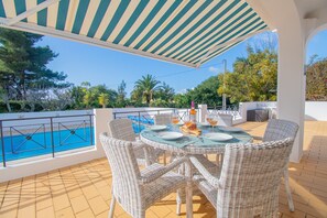 An outdoor dining area with wicker chairs and a table set for two, overlooking a pool and surrounded by greenery.