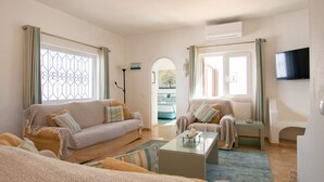 A living room with a sofa, armchairs, a coffee table, and a TV. There are windows with curtains, a door, and an air conditioning unit on the wall. - 61 vdm Superb Villa on Vale do Milho With Heated (Lagoa)