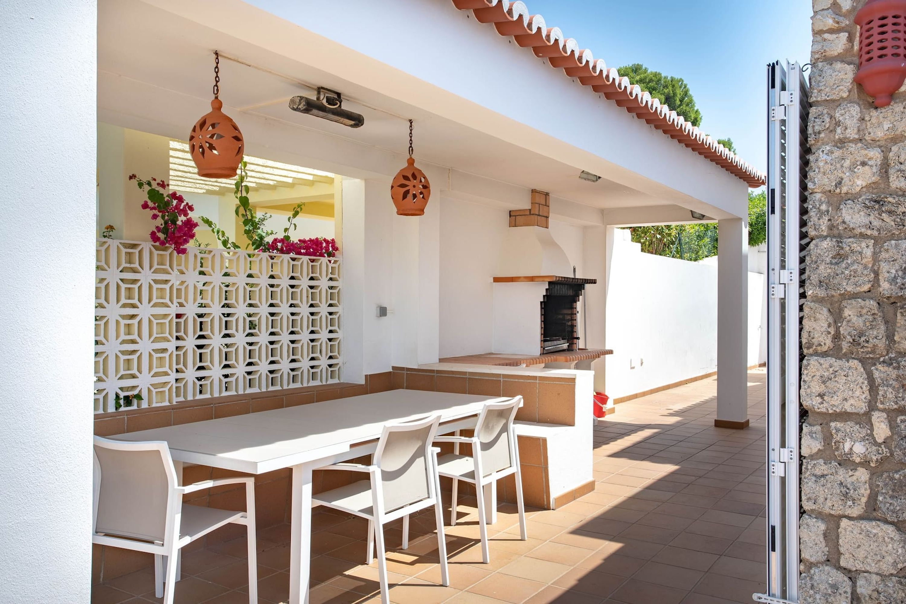 An outdoor patio with a white table and chairs, a decorative wall with geometric patterns, hanging terracotta lanterns, and a stone wall.
