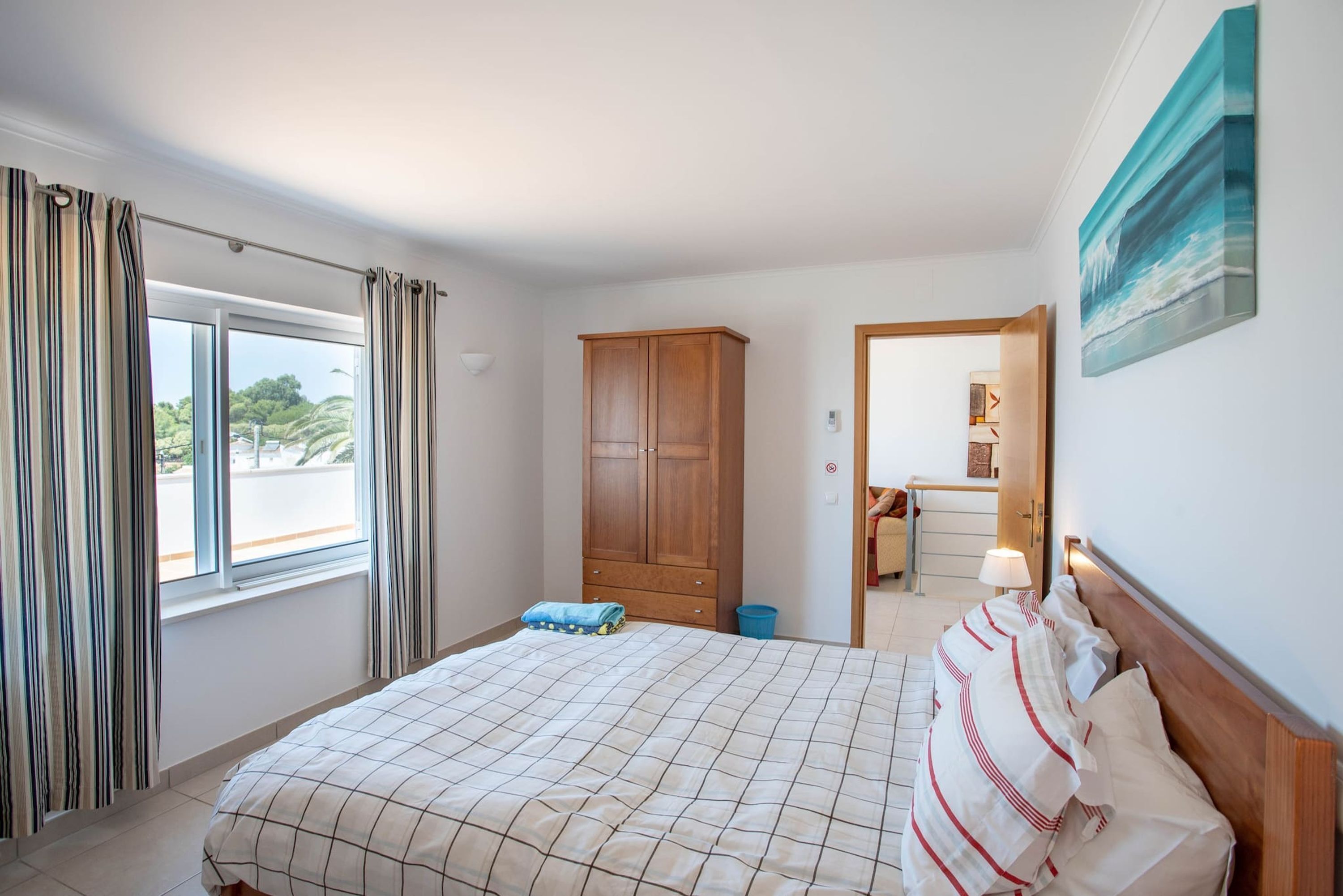 A bedroom with a bed, curtains, a wardrobe, a painting, and a view through the window.