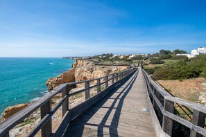 A wooden boardwalk leading to a coastal area with rocky cliffs and a clear blue sky. - C65 Quinta do Paraiso (Lagoa)