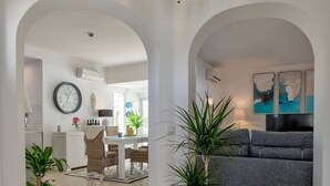 Ein modernes Wohnzimmer mit grauem Sofa, einem Essbereich mit weißem Tisch und einer Küche im Hintergrund.
