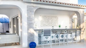 Ein moderner Poolbereich im Freien mit Whirlpool, Bar mit Sitzgelegenheiten und Blick auf ein weißes Gebäude mit Bögen und einem Ziegeldach.