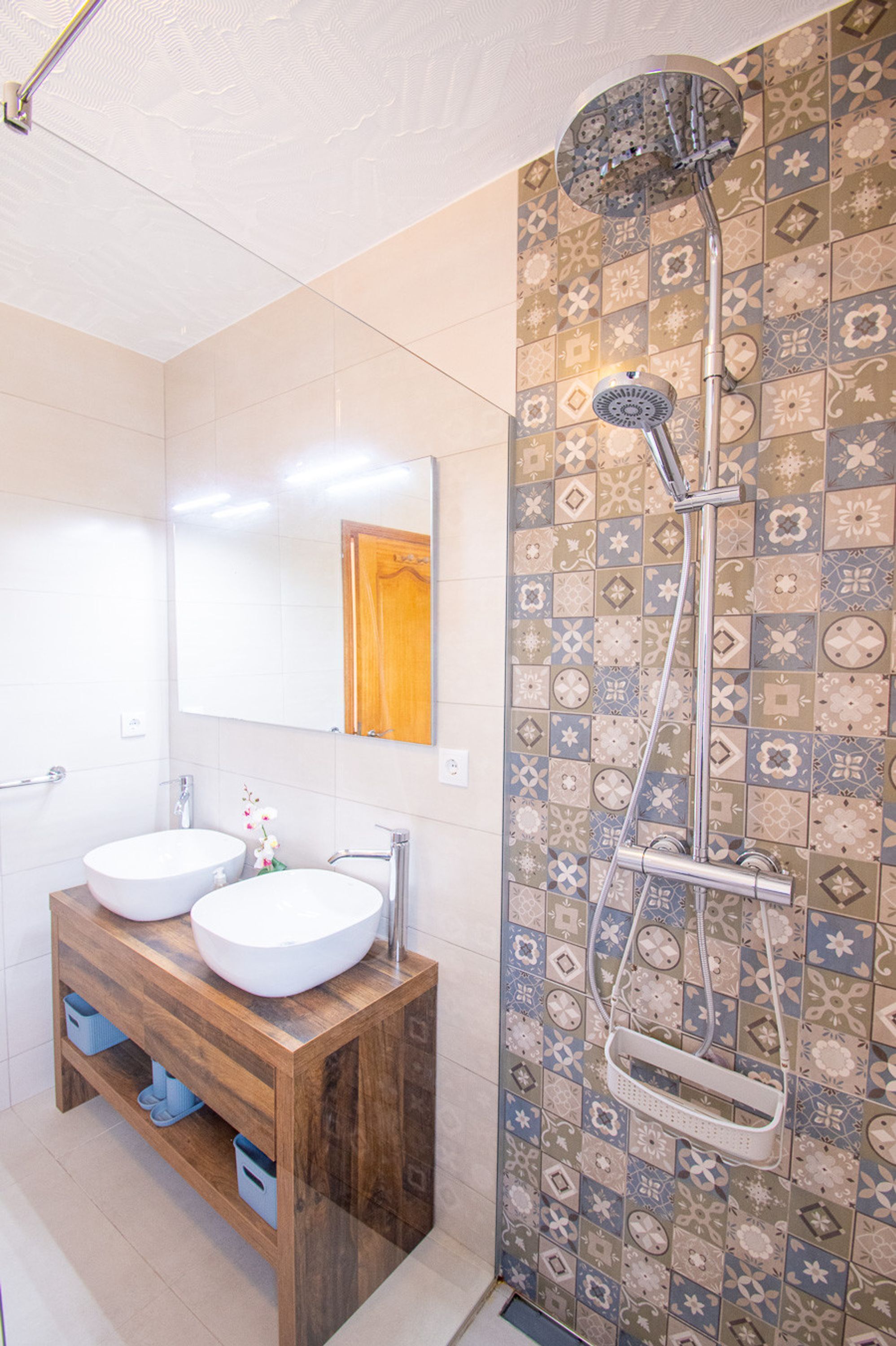 A bathroom with two sinks, a wooden vanity, a mirror, and a shower with a patterned tile wall.