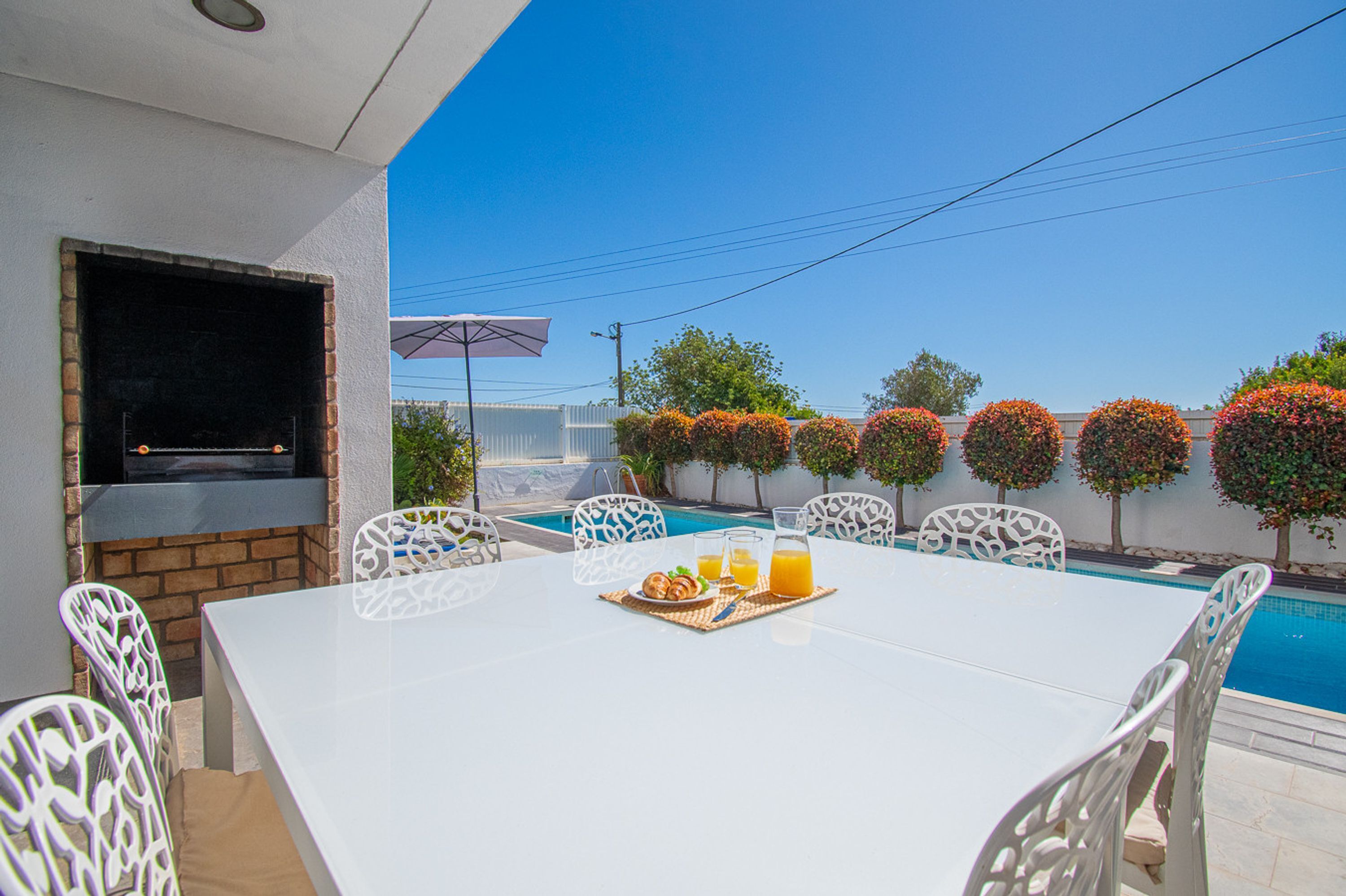 Une terrasse extérieure avec une table et des chaises blanches, un barbecue et une piscine.