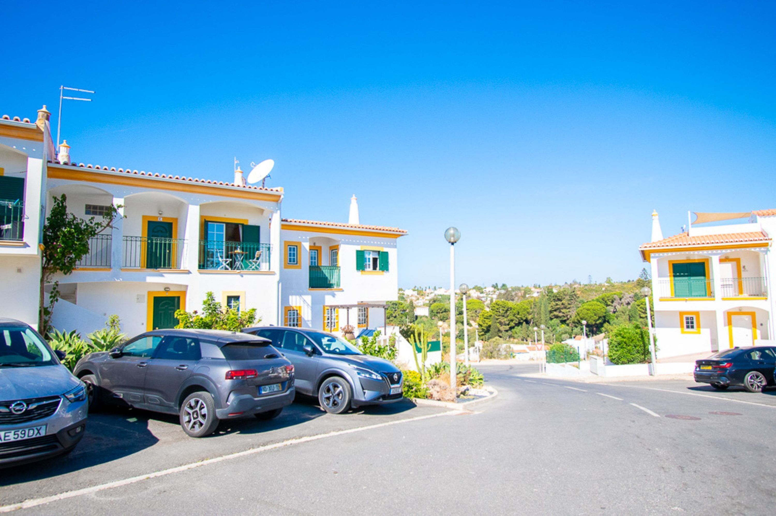 A row of white buildings with balconies, parked cars, and a clear blue sky.