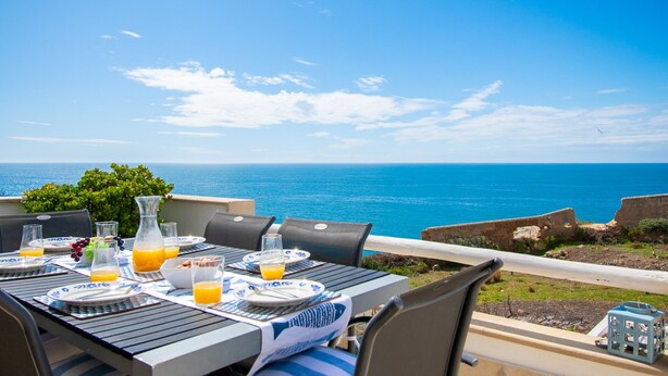 An outdoor dining table set with plates, glasses, and a pitcher of orange juice, overlooking a scenic ocean view. - Casa Mar a Vista Best View in Town (Lagoa)