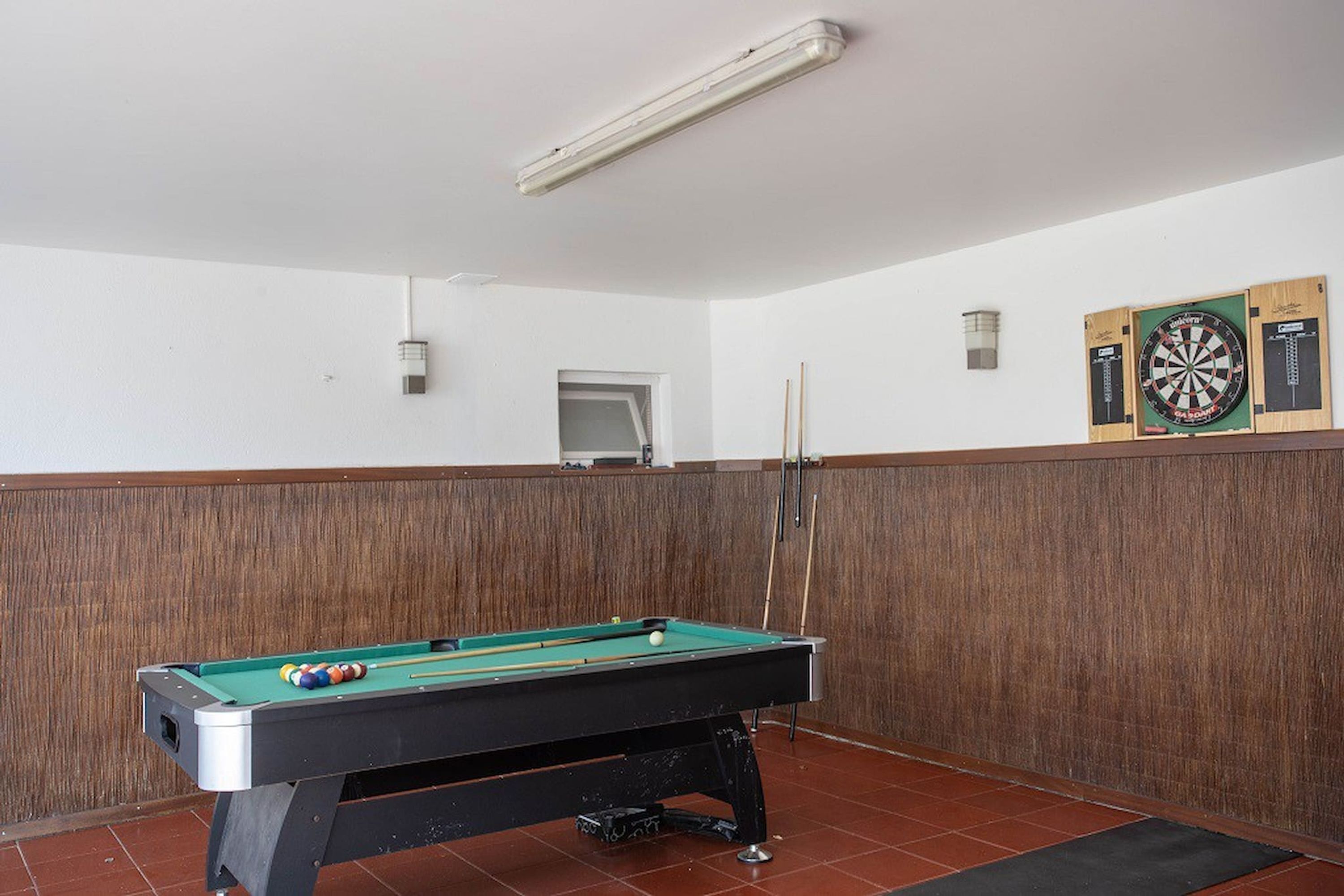 A pool table with balls and cues, a dartboard, and a small window in a room with wooden paneling and tiled flooring.