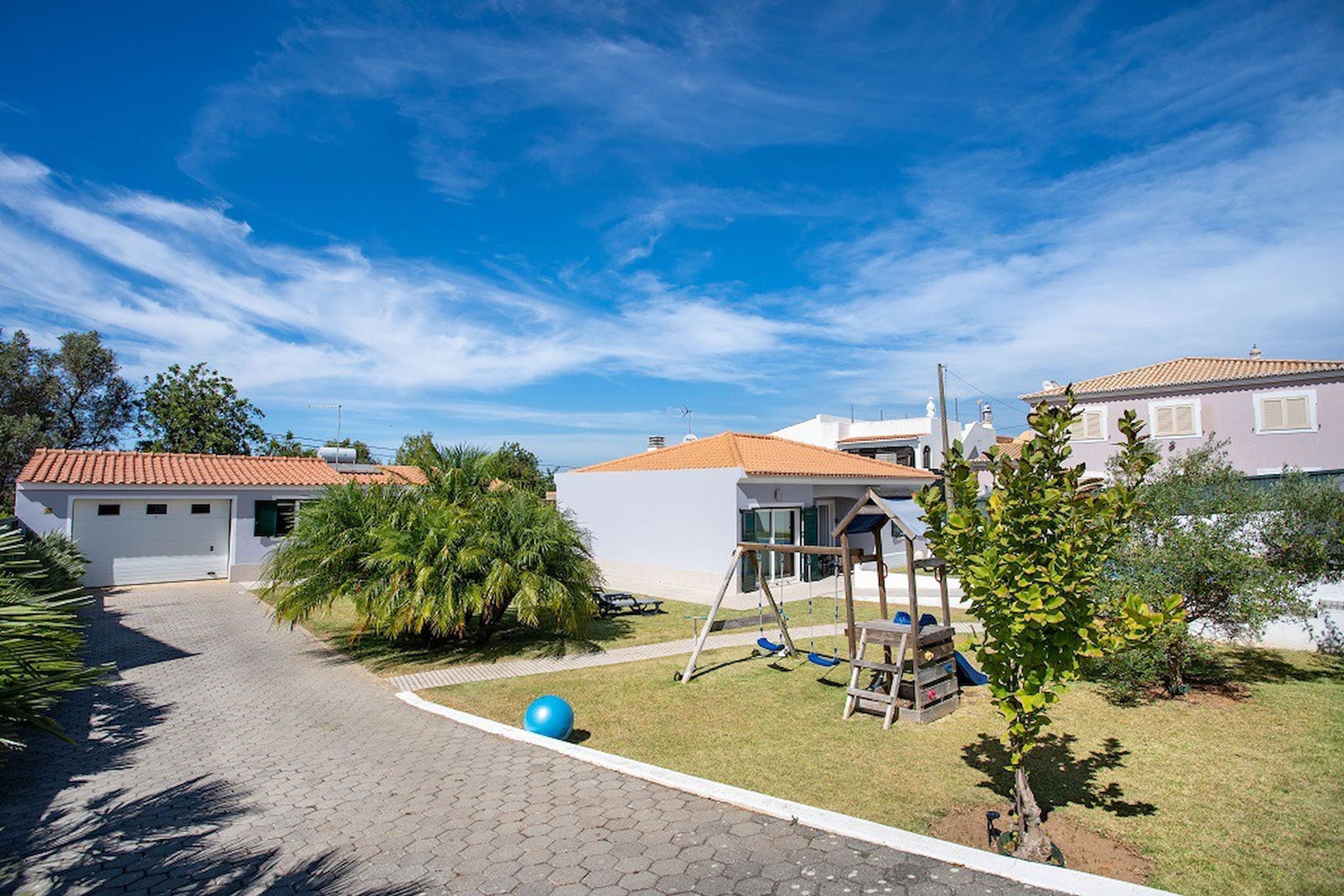 A residential area with a paved driveway, a swing set, and a small tree.