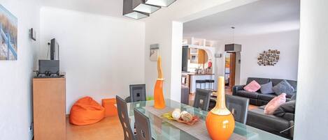 A modern living room with a glass dining table, orange bean bag chairs, and a sofa with cushions.