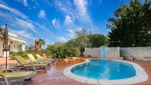 Una piscina con tumbonas, un muro de piedra y un cielo azul despejado.