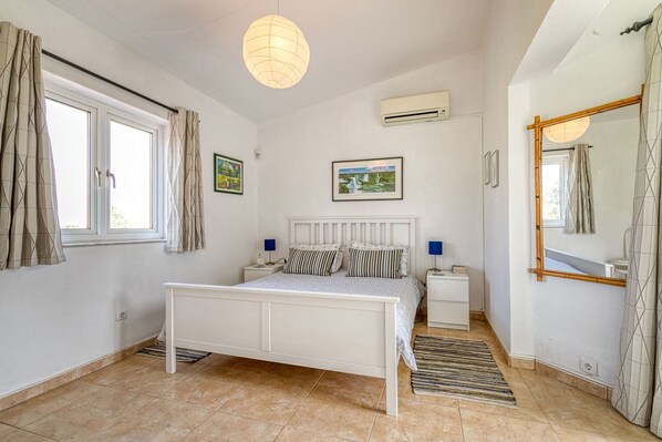 A bedroom with a bed, bedside tables, a hanging light fixture, a window with curtains, an air conditioning unit, and a mirror.