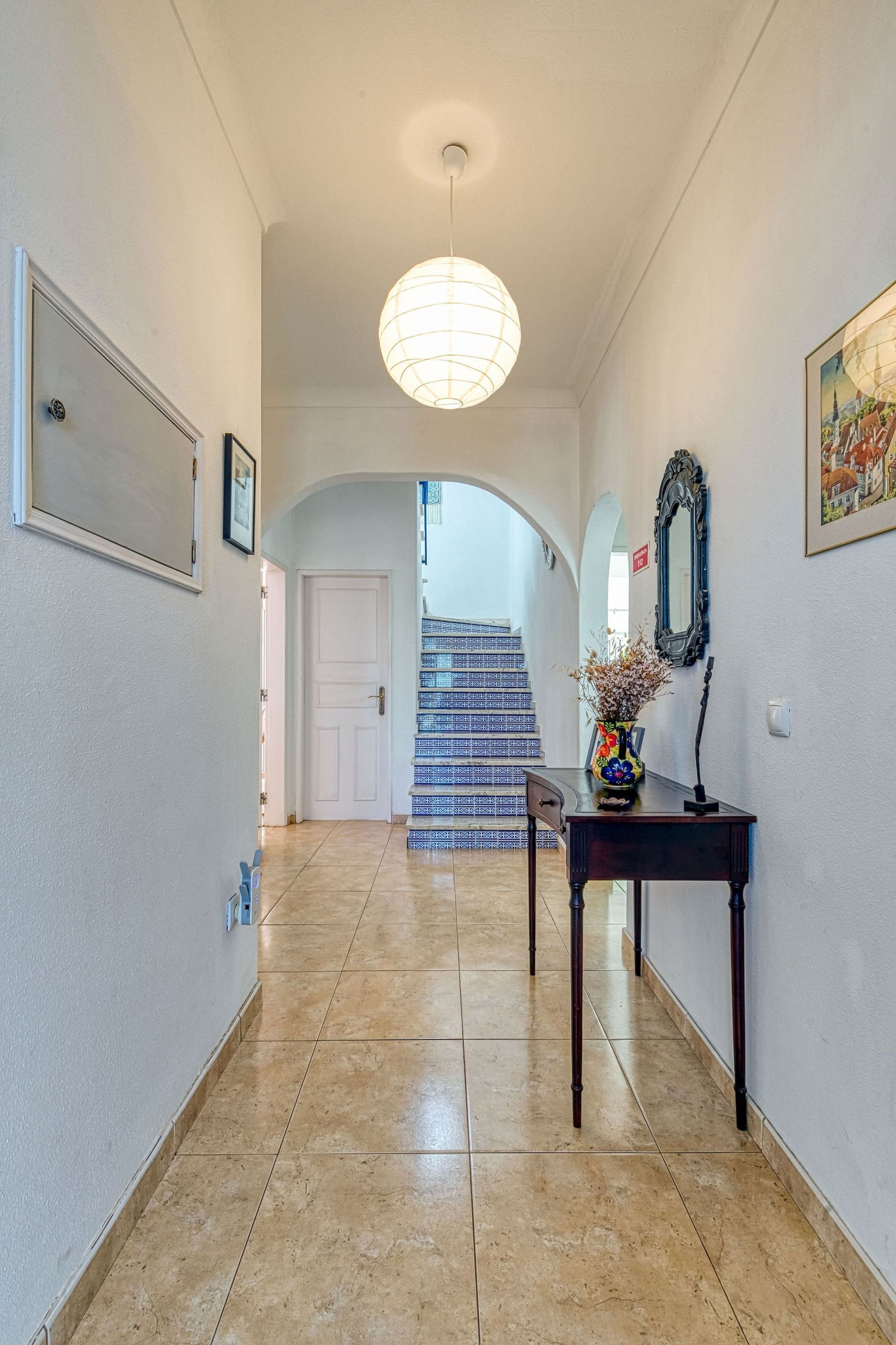 A hallway with a tiled floor, a staircase with patterned steps, a dark wooden console table with a vase, and a hanging light fixture.