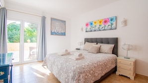 Un dormitorio con cama, mesita de noche, lámpara, un cuadro y vistas a través de la puerta de vidrio.