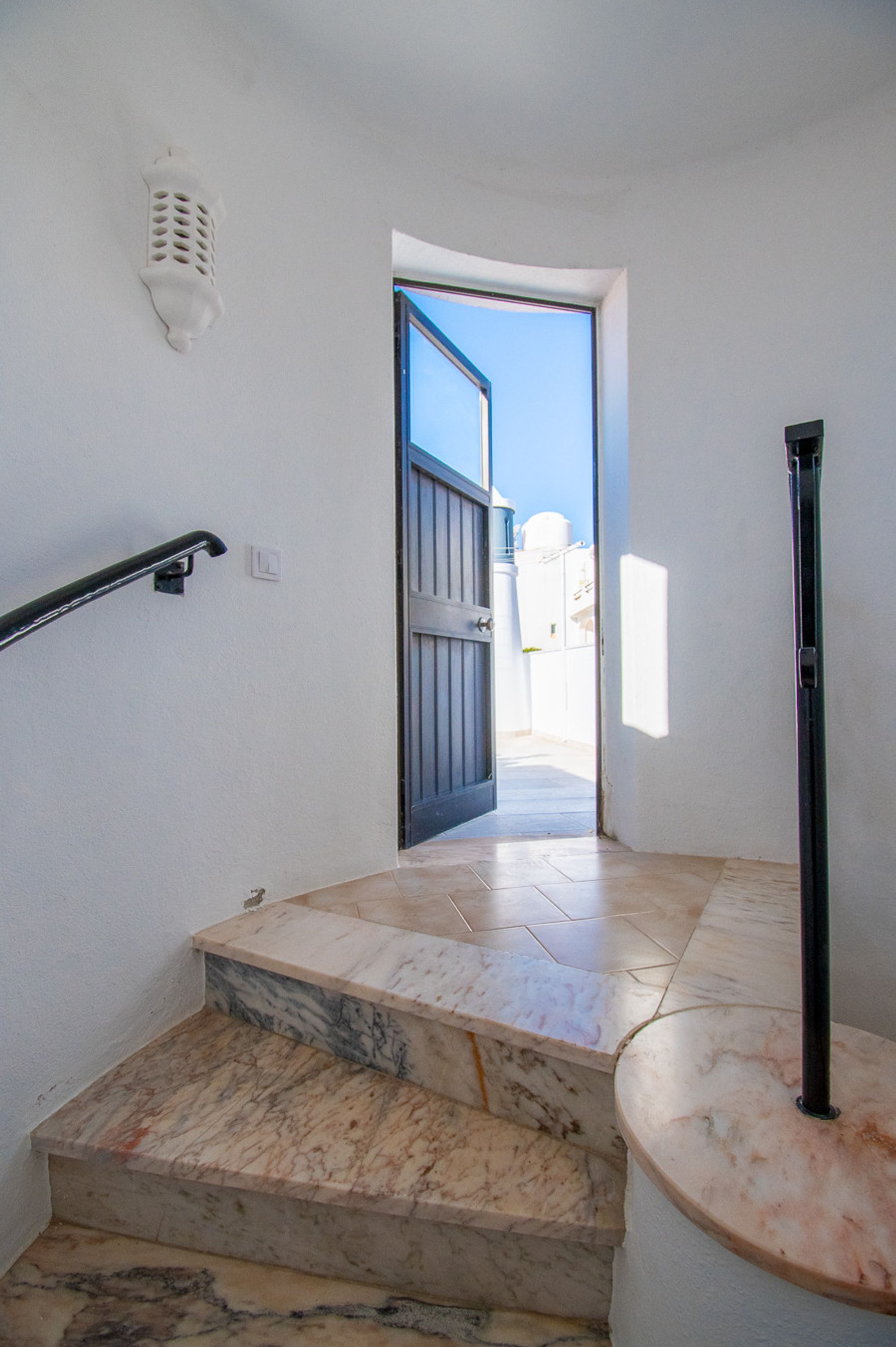 A staircase with marble steps leading to an open door, a handrail on the left, and a wall-mounted light fixture.