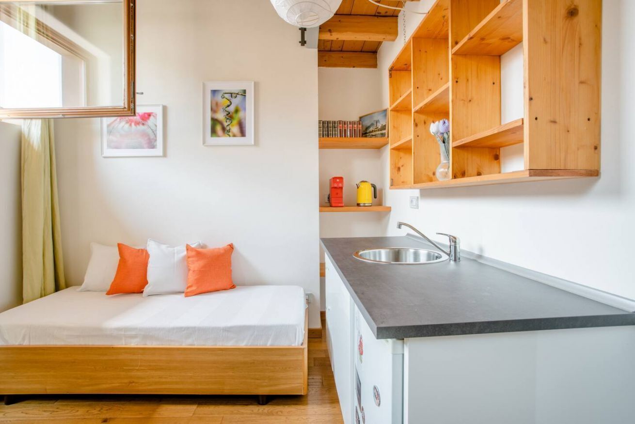 A bedroom with a wooden bed, a small kitchenette with a sink, and wooden shelves above the bed.