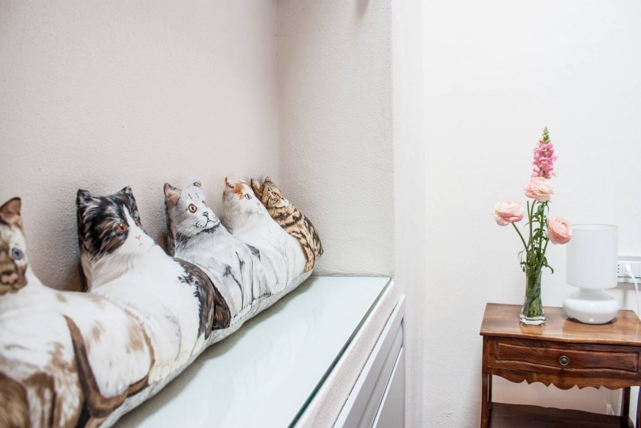 A corner of a room with a glass cabinet, three cat-shaped pillows, and a small wooden nightstand with a vase of flowers and a lamp.