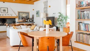 Amplia sala de estar con techo de madera, chimenea, mesa de comedor con sillas, estantería y televisor.