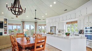 A spacious kitchen with white cabinetry, a large island with a sink, and a dining area with wooden chairs and a chandelier. - Paradise Found | Ocean & Marsh Views (Edisto Island)