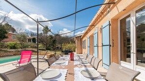 An outdoor dining area with a table set for six, surrounded by chairs, and a pool in the background.