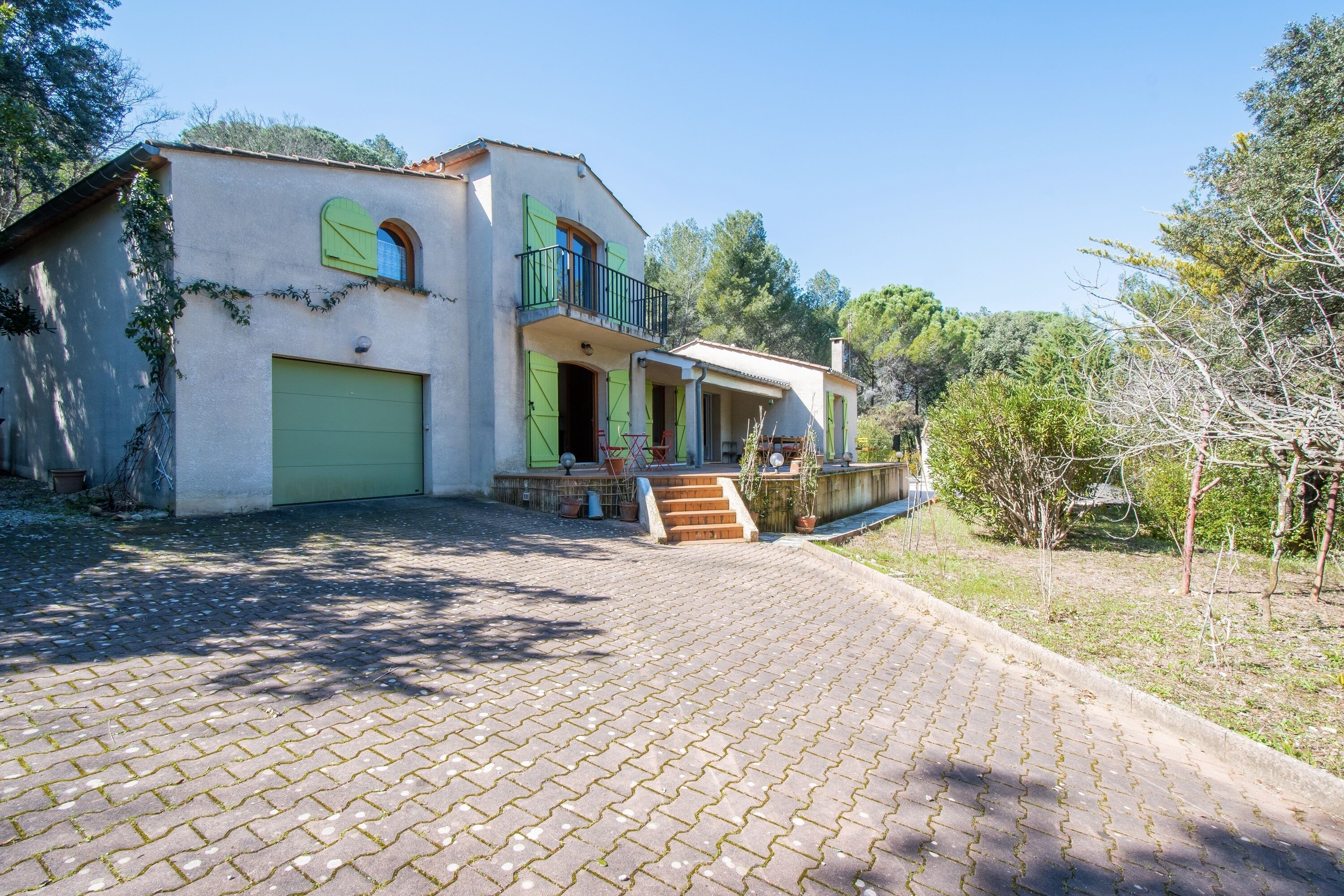 Een huis met twee verdiepingen, een groene garage, een balkon met een zwarte reling en een verharde oprit naar de ingang.