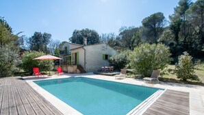 Una piscina rectangular amb un solàrium de fusta, envoltada de vegetació i una casa amb un balcó.