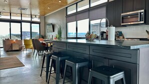 A modern kitchen with dark cabinetry, a large island with bar stools, a built-in microwave, and a view of the outdoors through large windows. - Cactus & Cabernet~Tesla Charger~Hot Tub~Waffle Bar (Quincy)