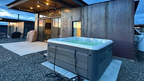 A modern outdoor hot tub area with a covered seating space and a building with large windows. - Cactus & Cabernet~Tesla Charger~Hot Tub~Waffle Bar (Quincy)