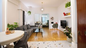 A modern living room with a sofa, a dining table, and a TV. There are plants and a bookshelf. - Ruby's Urban Residence Koukaki (Athens)