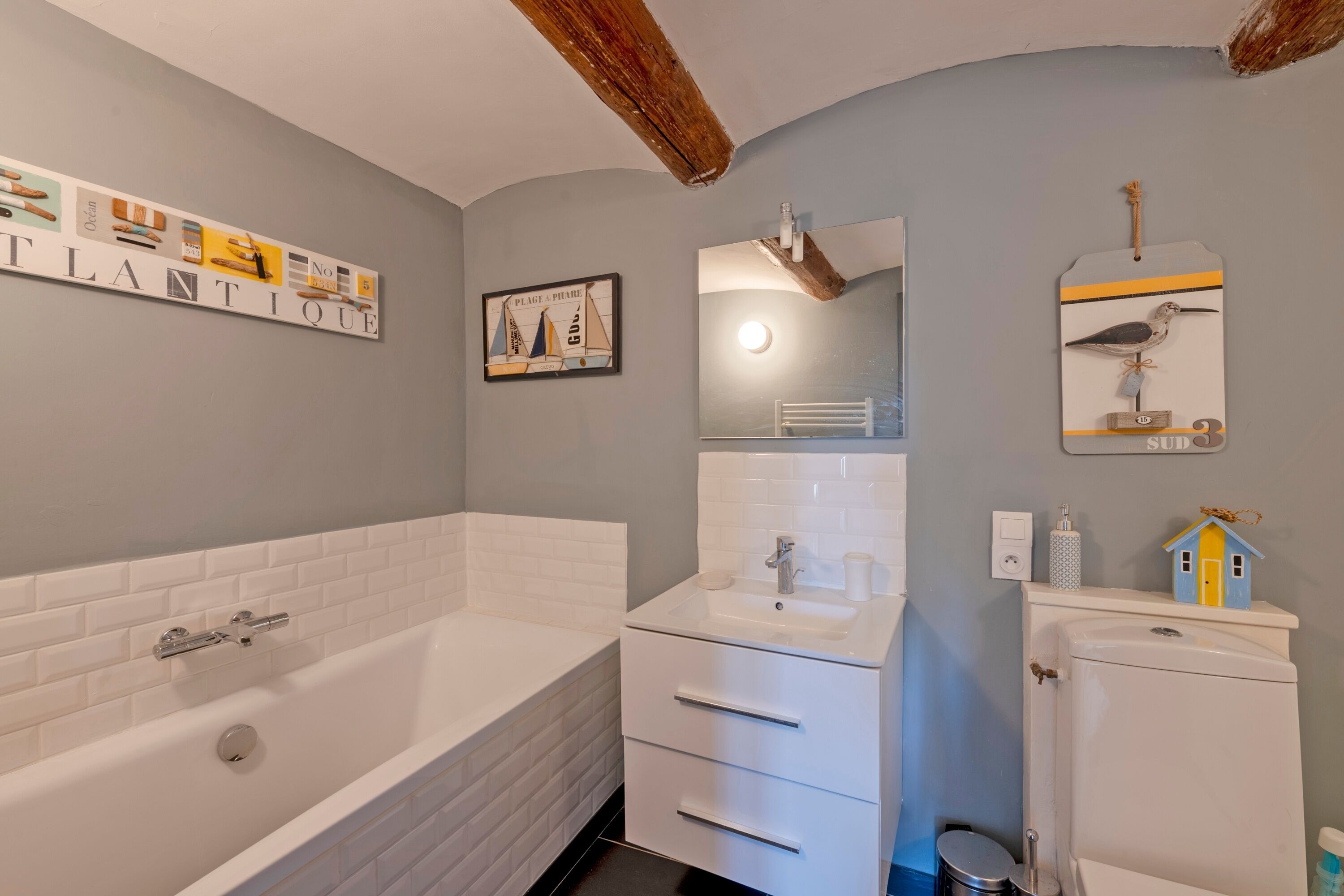 Ein Badezimmer mit einer weißen Badewanne, einem Waschbecken und einer Toilette. Über dem Waschbecken hängt ein Spiegel und an der Wand hängt ein gerahmtes Bild.