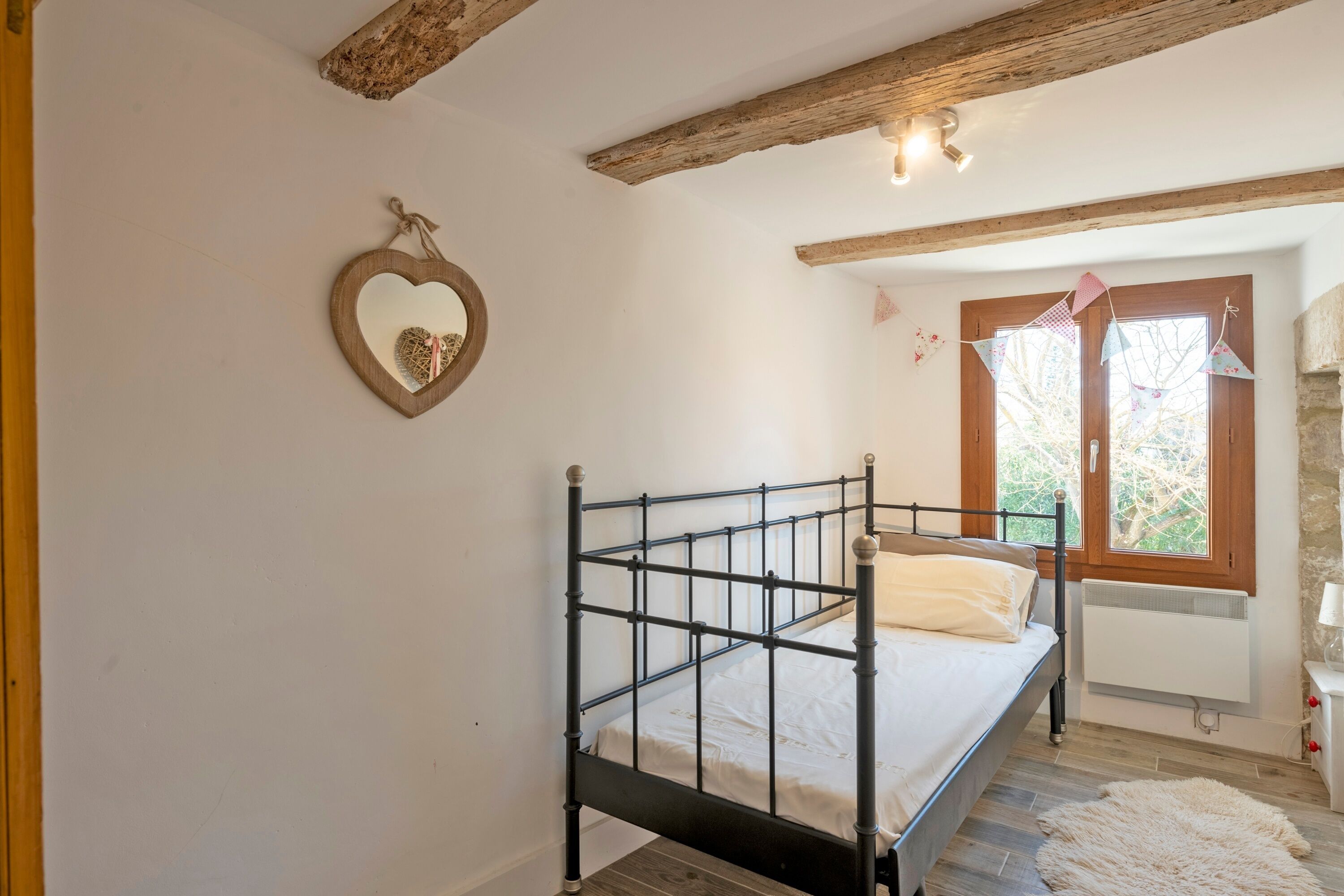 Un dormitorio con estructura de cama metálica, espejo con forma de corazón y ventana con vistas a árboles.