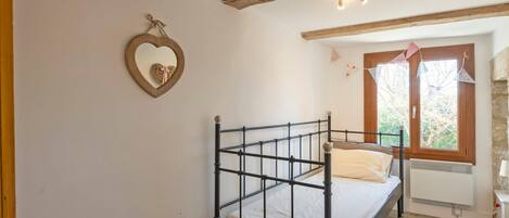 Un dormitorio con estructura de cama metálica, espejo con forma de corazón y ventana con vistas a árboles.