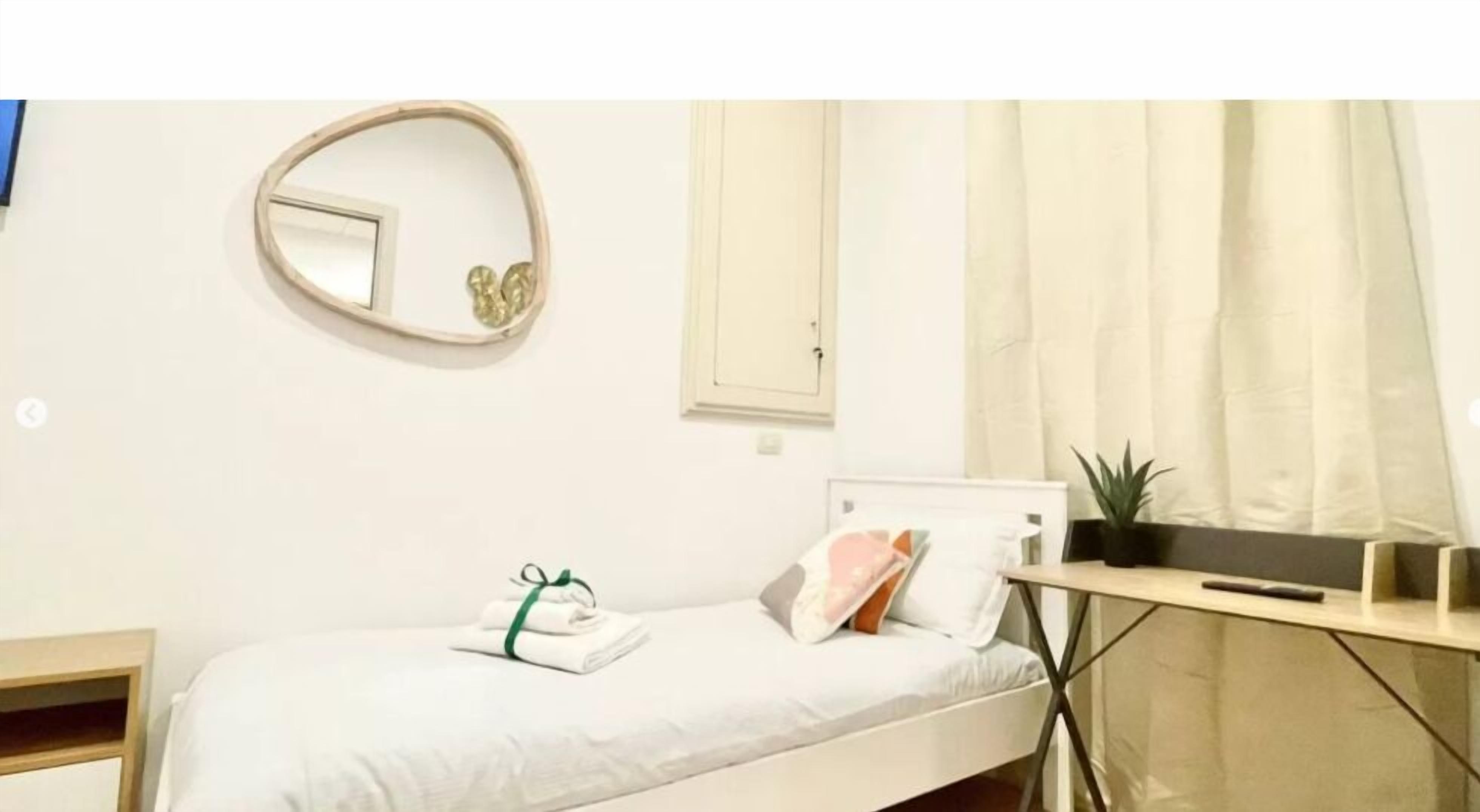 A neatly arranged bedroom with a bed, a bedside table, a mirror, a window with curtains, and a small potted plant.
