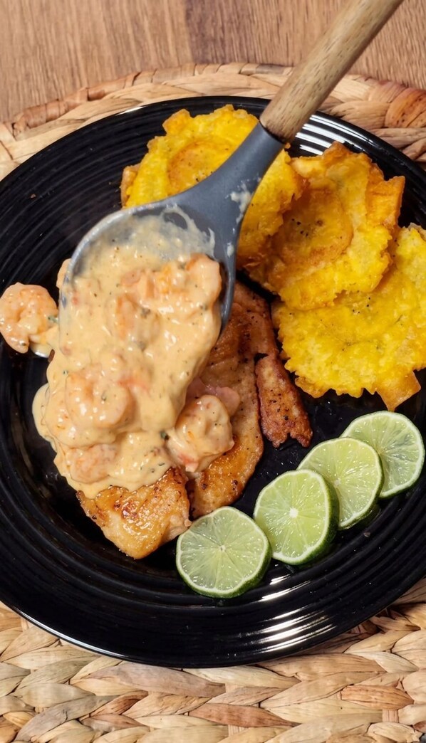 A black plate with grilled chicken, a creamy sauce, fried plantains, and lime wedges.