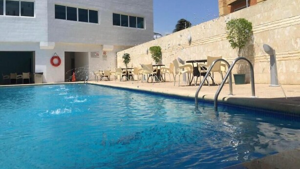 Une piscine avec des chaises longues et des tables, une bouée de sauvetage, et un bâtiment doté de nombreuses fenêtres et d’un escalier.
