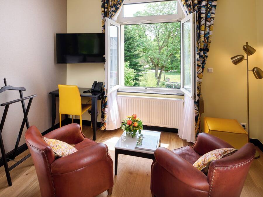 A room with two leather armchairs, a small table with a vase of flowers, a desk with a phone, a TV mounted on the wall, and a window with a view of trees.