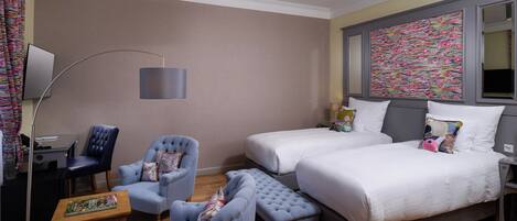 A hotel room with two beds, a desk, a chair, a lamp, and a colorful artwork on the wall.
