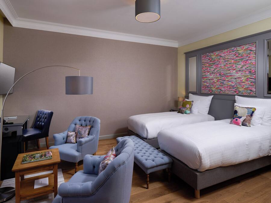 A hotel room with two beds, a desk, a chair, a lamp, and a colorful artwork on the wall.