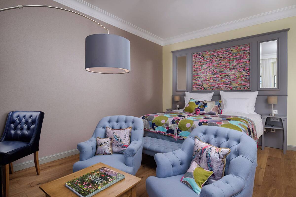 Un dormitorio con cama, dos sillones, una mesita de centro, una lámpara y una obra de arte colorida en la pared.