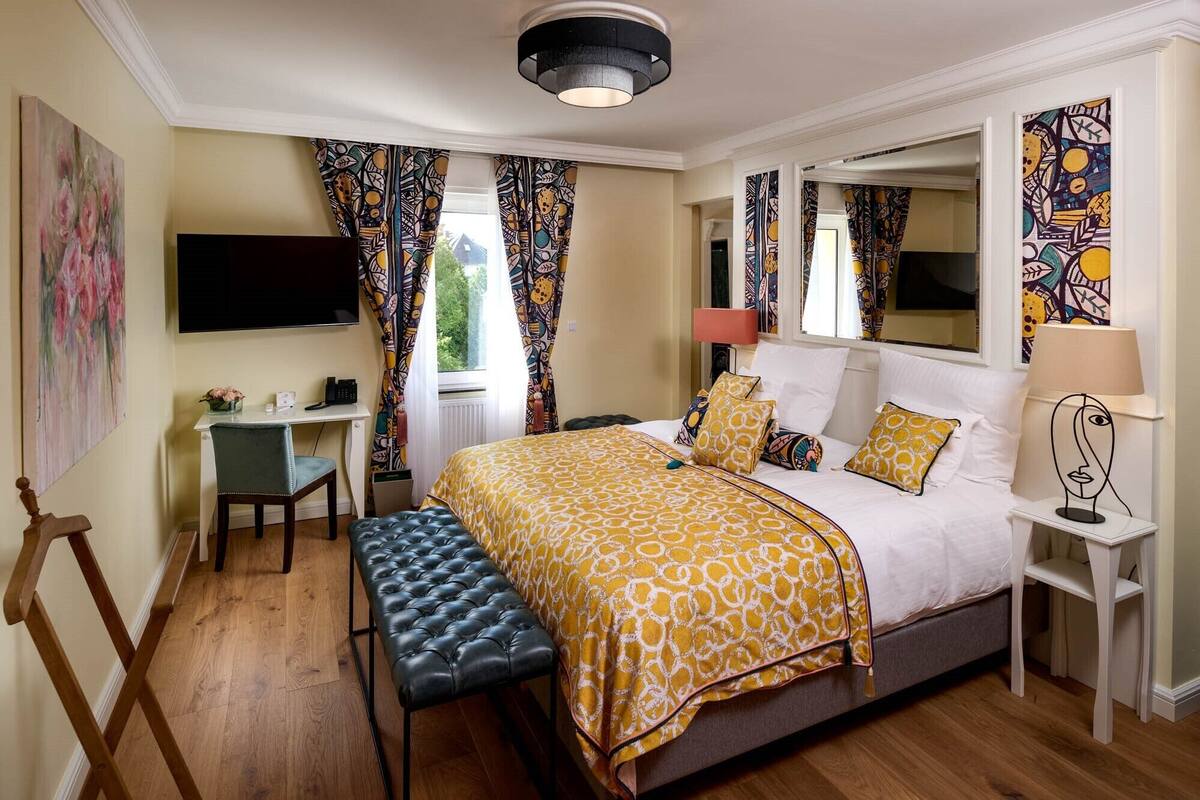 Un dormitorio con cama, escritorio, silla, banco, televisor, espejo y lámpara.