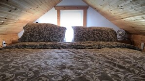 Ein Schlafzimmer mit schrägen Wänden, zwei gemusterte Kissen, ein Bett mit einer gemusterten Tagesdecke und ein Fenster mit Blick nach draußen.