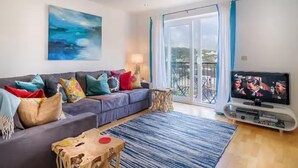 A living room with a grey sofa, colorful cushions, a large abstract painting, a TV, and a view of a balcony. - The Cove, Porthtowan (Porthtowan)