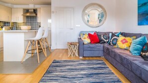 A modern living room with a grey sofa, colorful cushions, a round mirror, and a kitchen area with white cabinets and a dining set. - The Cove, Porthtowan (Porthtowan)